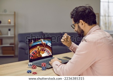 Young man gambling in virtual online casino on Internet. Happy guy sitting at desk table with poker chips, looking at casino wheel on screen of modern laptop computer and doing yes fist gesture Royalty-Free Stock Photo #2236385315
