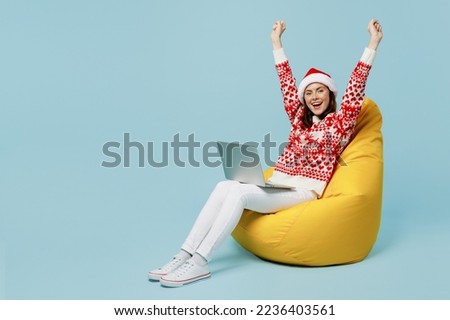 Full body young merry IT woman wear red sweater Santa hat posing sit in bag chair work hold use laptop pc computer do winner gesture isolated on plain blue cyan background. Happy New Year 2023 concept