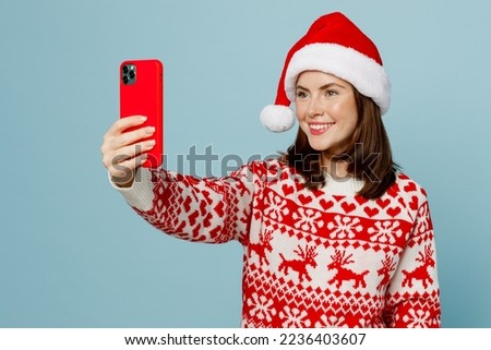 Young fun merry woman 20s wear warm red knitted sweater Santa hat posing doing selfie shot on mobile cell phone isolated on plain pastel light blue cyan background. Happy New Year 2023 holiday concept