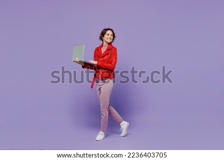 Full body young smiling happy woman 20s wear red leather jacket hold use work on laptop pc computer look aside isolated on plain pastel light purple background studio People lifestyle fashion concept