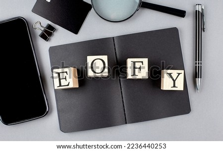 Word EOFY, End of Financial Year on a wooden block on black notebook with smartpone, credit card and magnifier