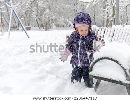 Child plays with snow in winter, little kid walks in snowy urban park during snowfall. Baby girl is on snow background. Theme of cold, frost, winter, season and weather.