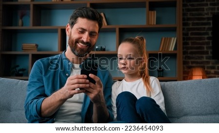 Cute small baby girl child resting on cozy couch with caring young father man playing game on smartphone watching media content on phone using mobile apps shopping online taking family selfie together