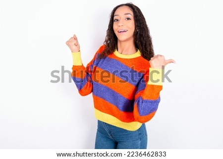Hooray cool Beautiful teen girl with curly hair wearing striped knitted sweater over white background point back empty space hand fist
