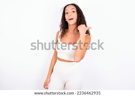 Impressed Beautiful teen girl with curly hair wearing white sport set over white background point back empty space