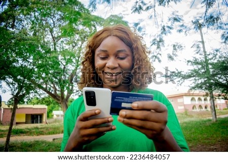 Satisfied lovely woman holds modern cell phone with credit card in school campus, technology concept, blurred picture