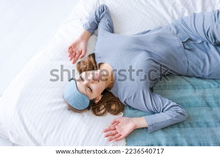 Relaxed joyful caucasian young woman stretches her arms after waking up, rejoices in a good day, enjoys a pleasant sleep, smiles sincerely, wears a sleep mask and pajamas, with a happy expression