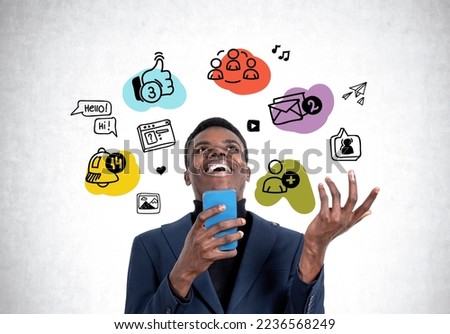 Laughing black businessman with smartphone in hands, looking up at colorful social media doodle icons on grey concrete wall background. Concept of chatting