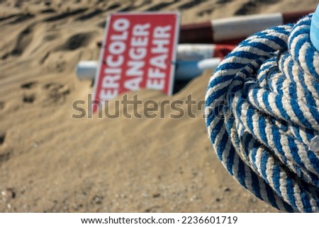 In the fine sand on the sea beach lies a sign with the inscription "Danger" in three different languages. A rope for the barrier is next to it.