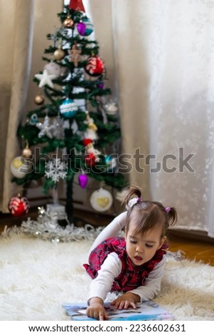 little girl in a red dress with ponytails is reading a book on floor in front of decorative Christmas tree at home. Little beautiful girl reads a fairy tale. Family holiday.