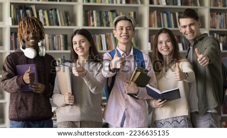 Happy multiethnic first year college students posing in university library, looking at camera, standing at bookshelves, showing like thumb up gestures, smiling, laughing, recommending education