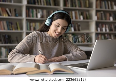 Happy Latin student girl in wireless earphones studying in public library, writing notes at laptop, open books, listening music for focus, watching learning class, webinar, lesson, making summary