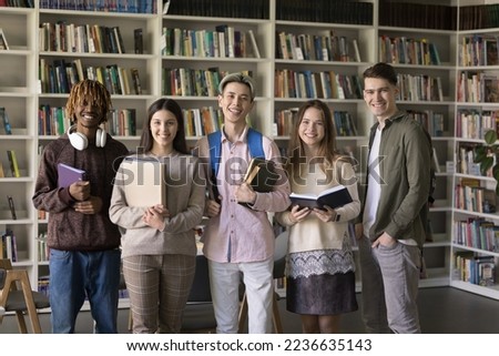 Happy diverse college students posing for group portrait in library, standing together in row at bookshelves, holding textbooks, studying books, looking at camera, smiling