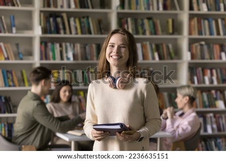 Happy pretty Caucasian student girl with headphones on neck visiting university library for work on study project, holding copybook, notebook, looking at camera, smiling, laughing, enjoying education
