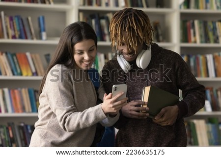 Diverse couple of happy classmates, college students using smartphone in library together. Latin high school girl showing learning video on cellphone screen to African friend, laughing, smiling Royalty-Free Stock Photo #2236635175