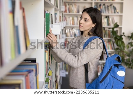 Focused pretty college student girl with backpack taking textbooks from shelf in university library, enjoying studying, literature hobby, reading, touching covers. Bookshop customer picking books