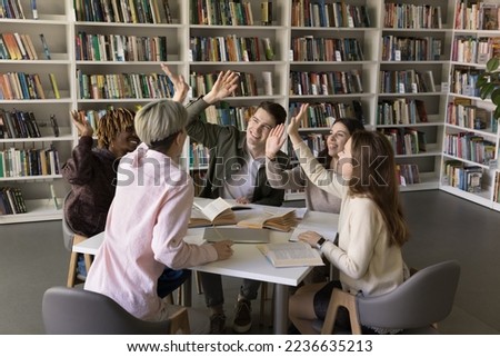 Diverse group of excited students enjoying teamwork, education process in college library, giving team high five, clapping hands, laughing, sitting together at table with open books