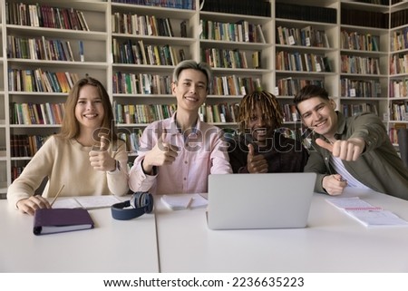 Diverse group of happy fresh college students enjoying teamwork in library, sitting at table together, smiling, laughing, showing thumb up, like hand gesture at camera, promoting education