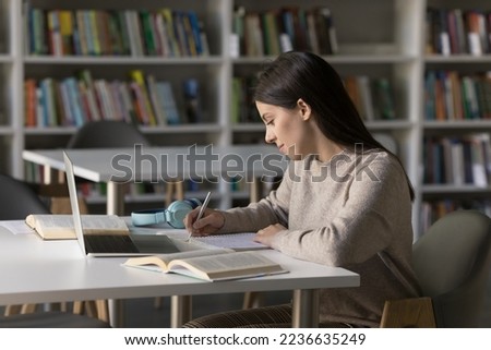 Focused pretty student girl studying in public library, writing draft, notes, article, essay, webinar summary, working, sitting at desk with open books, laptop computer. Side view Royalty-Free Stock Photo #2236635249