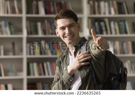 Cheerful satisfied handsome student guy recommending college, university education, giving positive feedback, studying in library, showing like hand sign at camera, making thumb up, smiling at camera