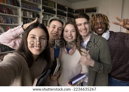 Team of cheerful multiethnic teen students taking group selfie in university library, showing thumb up like hand, making peace gesture, smiling, laughing, looking at camera for self portrait