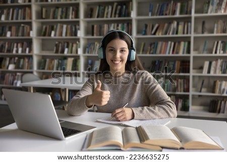 Happy confident student girl in headphones showing hand like sign, thumb up gesture, looking at camera, smiling, working in university library, writing essay, article. Head shot portrait