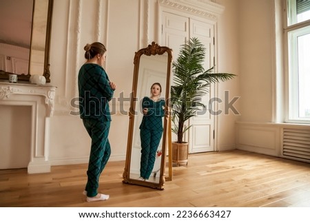 Lonely female looking at mirror in her reflection in living room at home. Alone middle aged adult woman standing and looks in mirror. Concept of adult domestic life and depression. Copy text space Royalty-Free Stock Photo #2236663427