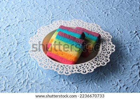 Rainbow cake on white plate with blue background. The background have textured. Cake Pelangi