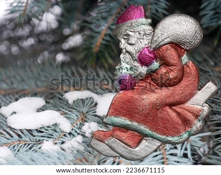 Santa Claus in red fur coat, felt boots and mittens with bag of gifts. Rides on sled from mountain. Vintage Christmas tree toy on background of spruce branches with snow.  Focus on foreground.
