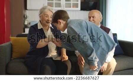 The little boy visited his grandfather and celebrated Eid al-Fitr by kissing their hands. Happy senior couple sitting on sofa kissing and hugging their little grandchild.  Royalty-Free Stock Photo #2236687627