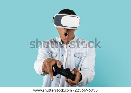 Glad young african american guy in casual and vr glasses with open mouth and joystick has fun with technology, plays online game, isolated on blue background, studio. Virtual world and entertainment