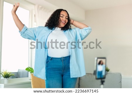 African american plus size female influencer recording video on smartphone, dancing on camera at home. Positive lifestyle blogger making content for her blog, filming for social media