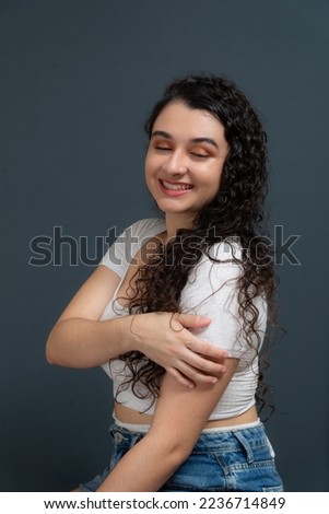 Cheerful beautiful young woman with hand on shoulder wearing white shirt. Against gray background. concept of youth