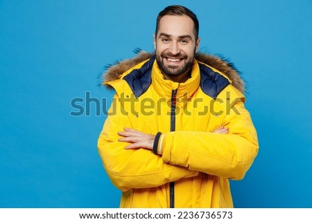 Young smiling happy fun cheerful cool caucasian man 20s wearing yellow down jacket hold hands crossed folded isolated on plain blue color background studio portrait. People winter lifestyle concept