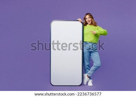 Full body young smiling fun woman 30s she wear casual green knitted sweater stand point finger on big huge blank screen mobile cell phone with area isolated on plain pastel purple background studio