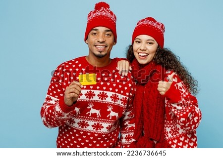 Merry fun young couple two man woman wear red Christmas sweater Santa hat posing hold credit bank card show thumb up isolated on plain pastel light blue background. Happy New Year 2023 holiday concept