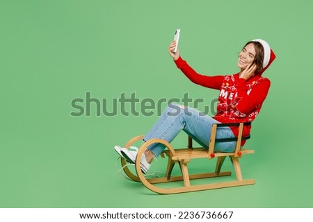 Full body merry young woman wear xmas sweater Santa hat posing sit on sleigh sledding doing selfie shot on mobile cell phone isolated on plain pastel light green background Happy New Year 2023 concept