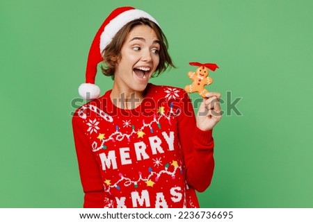 Merry surprised young woman wear knitted xmas sweater Santa hat posing hol in hand traditional ginger cookie isolated on plain pastel green background. Happy New Year 2023 celebration holiday concept