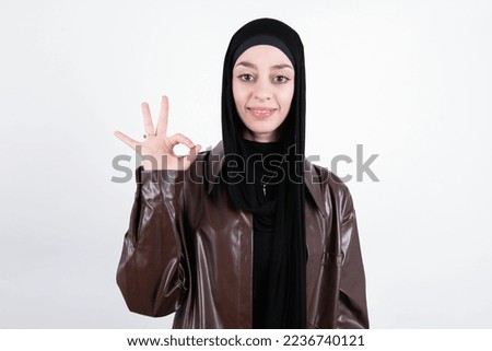 Glad attractive beautiful muslim woman wearing hijab and brown leather jacket over white background shows ok sign with hand as expresses approval, has cheerful expression, being optimistic.