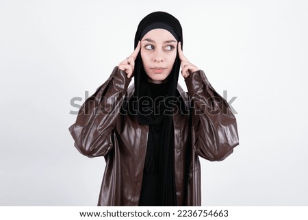 beautiful muslim woman wearing hijab and brown leather jacket over white background with thoughtful expression, looks away, keeps hand near face, thinks about something pleasant.