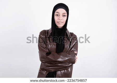beautiful muslim woman wearing hijab and brown leather jacket over white background bitting his mouth and looking worried and scared crossing arms, worry and doubt.