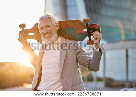 Smiling happy cool grey haired bearded older senior business man skater wearing suit holding skateboard standing in city on sunset outdoors after work. Old people freedom spirit concept. Portrait Royalty-Free Stock Photo #2236758871