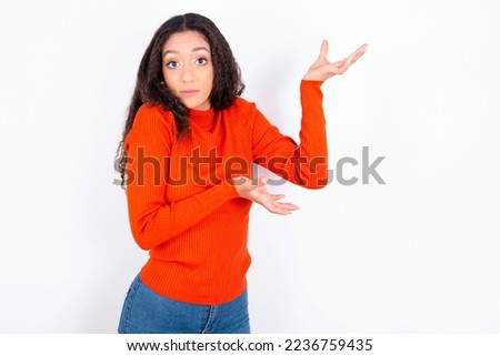 beautiful teen girl wearing knitted red sweater over white background pointing aside with both hands showing something strange and saying: I don't know what is this. Advertisement concept.