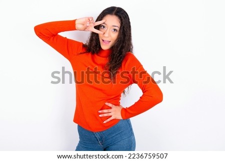 beautiful teen girl wearing knitted red sweater over white background making v-sign near eyes. Leisure, coquettish, celebration, and flirt concept.