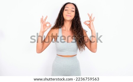teen girl with curly hair wearing grey sport set over white background relax and smiling with eyes closed doing meditation gesture with fingers. Yoga concept. Royalty-Free Stock Photo #2236759563