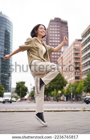 Vertical shot of young asian woman posing happy, raising hands up and dancing, triumphing, celebrating victory, enjoying day out in city. Royalty-Free Stock Photo #2236765817