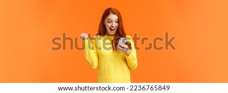 Girl received thousand likes under new picture. Cheerful and triumphing pretty redhead female fist pump say yes, cheering or celebrating good news, looking smartphone screen, orange background.