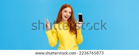 Positive and excited, charming redhead woman in yellow sweater, showing smartphone screen, advertise online store, mobile application, make peace sign and smiling joyfully, blue background.