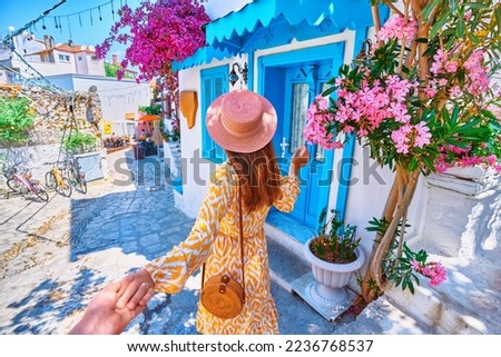 Follow me concept. Girl traveler wearing dress and hat walks on beautiful colorful flower street with white houses and blue doors in a European city Royalty-Free Stock Photo #2236768537