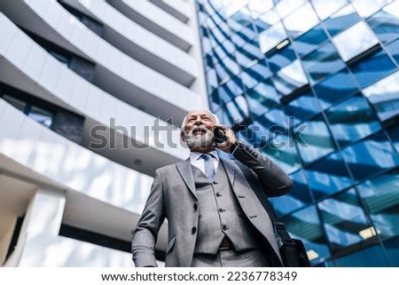 Low angle view of senior businessman talking on smart phone. Elderly professional wearing full suit is carrying laptop bag. He is standing against business office building. Royalty-Free Stock Photo #2236778349
